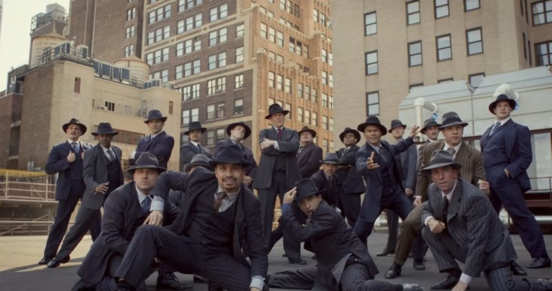 A large group of men in suits and old-fashioned fedoras are in front of a large New York building. Some stand while others kneel.